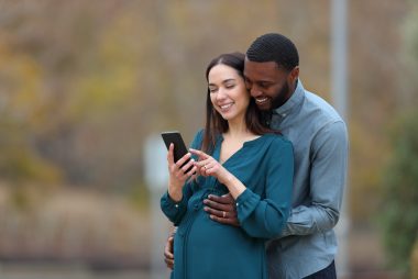 Interracial couple with a pregnant wife and her husband checking smart phone in a park